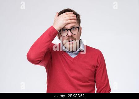 Giovane depresso e frustrato in maglione rosso che tocca la testa con la mano mentre si trova su sfondo bianco. Egli falsare il suo pin della carta di credito Foto Stock