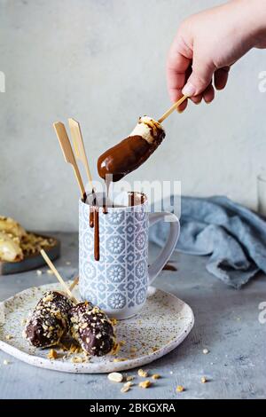 Banana congelata su un bastone ricoperto di cioccolato fondente e briciole di noci. Un dessert veloce e delizioso per i bambini, per un compleanno, per un bar di caramelle. Vegano d Foto Stock