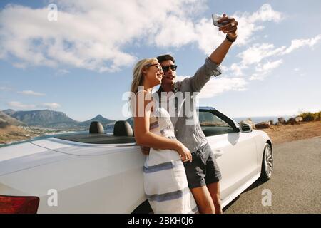 Bella coppia che scatta foto di se stessi appoggiandosi contro la loro cabriolet. Coppia che si gode un viaggio in auto. Foto Stock