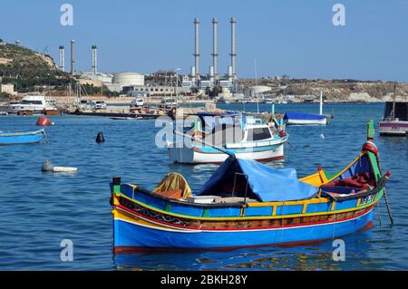 Barche ormeggiate nel porto del villaggio di pescatori di Marsaxlokk, Malta. La centrale elettrica di Delimara è raffigurata sullo sfondo. Foto Stock