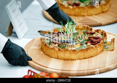 Mani di un cameriere in guanti neri mettendo sopra torte con germogli, tritate a pezzi con pinze di metallo sul tavolo del banchetto dove ci sono già Foto Stock