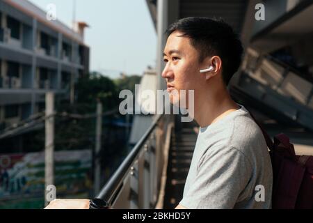 Primo piano di un giovane che ascolta musica con i cuscinetti wireless mentre si è in treno alla stazione. Un ragazzo asiatico che ascolta musica in movimento. Foto Stock