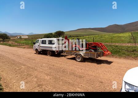 Overberg, Caledon, Capo Occidentale, Sud Africa. 2019. Veicolo agricolo che traina un rimorchio che trasporta una macchina scavatrice mini lungo una strada sterrata. Foto Stock