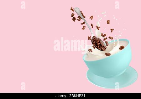 Lettere di cacao di cereali che cadono in ciotola blu con spruzzare latte su fondo rosa. Colazione a secco per bambini. Spazio di copia. Foto Stock