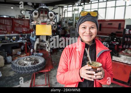 Ritratto di donna che beve tè compagno in officina meccanica moto Foto Stock