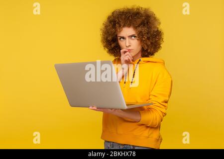 Confusa e sconcertata donna pensata con capelli ricci morbidi in stile urbano con cappuccio che tiene il laptop pensando a domande difficili, soluzione di riflessione. ind Foto Stock