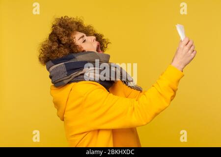 Influenza stagionale. Vista laterale della donna malata di influenza con capelli ricci morbidi, avvolti in sciarpa calda, starnuti in tovagliolo, sensazione di malessere, tosse e blowin Foto Stock