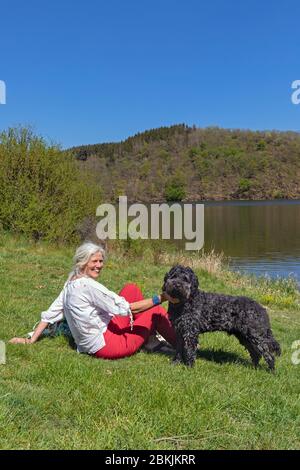 Europa, Lussemburgo, Insenborn, attraente donna anziana seduta sulla riva del Lac Sûre con il suo cane d'acqua portoghese Foto Stock