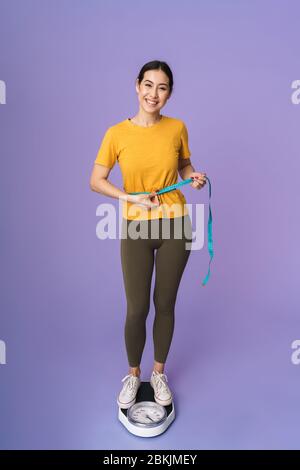 Tutta la lunghezza di una giovane allegra bella sportswoman in piedi su scale isolate su sfondo viola, tenendo nastro di misurazione intorno alla sua vita Foto Stock