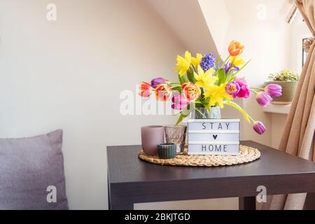 Tutto per un soggiorno tranquillo a casa. Scatola luminosa con messaggio di soggiorno a casa, bouquet di fiori freschi di primavera e candele sul tavolino da caffè, e cuscini sitti Foto Stock