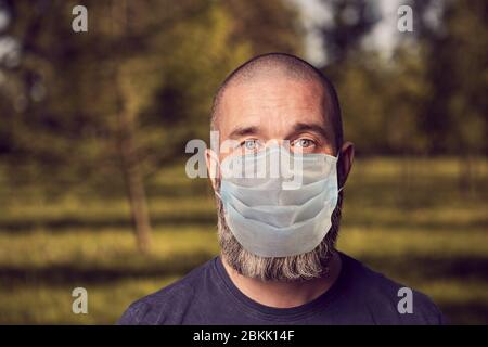 Uomo caucasico barbuto in una maschera protettiva sul viso sullo sfondo di un parco pubblico in una giornata estiva di sole durante la pandemia Covid-19. Un male bianco di mezza età Foto Stock