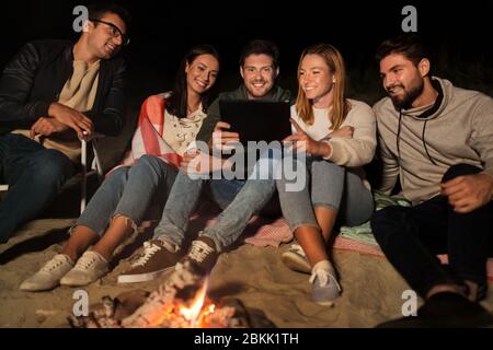 amici con un tablet pc al fuoco sulla spiaggia di notte Foto Stock