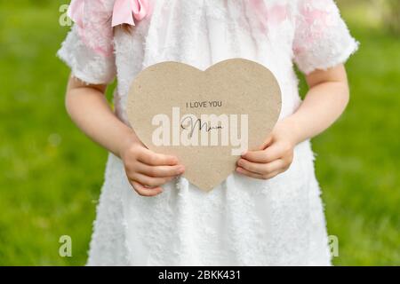 Ti amo mamma scritto su una carta a forma di cuore tenuto da una ragazza su sfondo d'erba Foto Stock