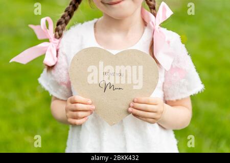 Ti amo mamma scritto su una carta a forma di cuore tenuto da una ragazza su sfondo d'erba Foto Stock