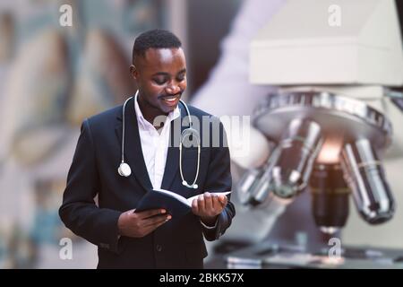 Medico africano con stetoscopio che indossa la tuta classica lettura di un libro vicino alla costruzione moderna clinica ospedale Foto Stock