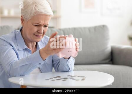 Crisi finanziaria. Donna Sad Senior che scuote Piggy Bank, prendendo le monete Foto Stock