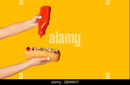 Primo piano di donna giovane che spreme ketchup su hot dog contro sfondo arancione, spazio copia. Panorama Foto Stock