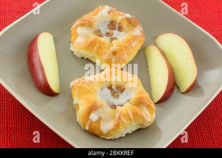 Due mini pasticcini danesi alla frutta di mela con glassa bianca e deliziose fette di mela rosse su piatto marrone taupe su tovaglia rossa Foto Stock