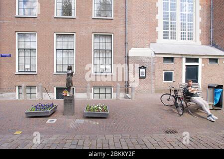 Un quotidiano locale di lettura vicino ai fiori, deposto alla statua di Anna Frank (1929-1945) vicino alla casa di Anna Frank, in una strada vuota durante la memoria Foto Stock