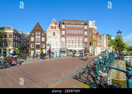 Biciclette parcheggiate lungo un canale vicino alla Chiesa Westerkerk, mentre i turisti e la gente del posto si godono i negozi e i ponti nel centro storico di Amsterdam Nether Foto Stock