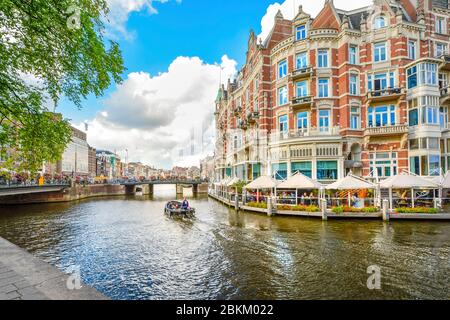 Una barca scivola passato a waterfront hotel e il cafe verso un ponte su uno dei canali principali della cittã , vicino al quartiere dei musei di Amsterdam, Paesi Bassi. Foto Stock