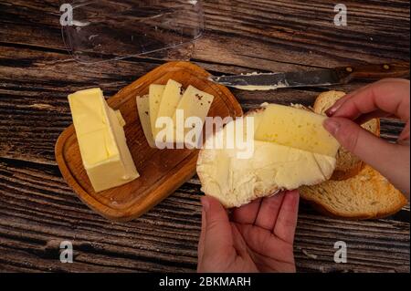 Qualcuno mette una fetta di formaggio su pane tostato di grano fresco con burro e un pezzo di burro in un piatto di legno su uno sfondo di legno. Primo piano Foto Stock