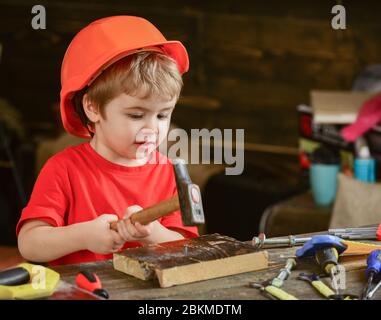 Piccolo ragazzo martellare chiodo in legno asse. Adorabile ragazzo seduto al tavolo da lavoro polveroso. Bambino concentrato che lavora con gli attrezzi. Foto Stock