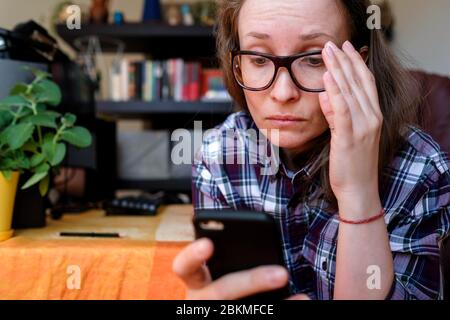 Una giovane donna sta leggendo dal telefono. Allontanare gli occhiali dal viso. Tiene un gadget in mano. Abiti da casa, lavoro a casa. Foto Stock