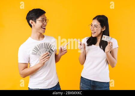 Immagine di coppia multinazionale eccitato con dollari e carta di credito puntando le dita l'una all'altra isolato su sfondo giallo Foto Stock