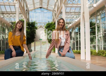 Due giovani donne caucasiche godono seduto sul bordo di una fontana con acqua all'interno di una serra luminosa Foto Stock