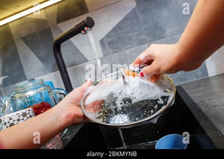 Mani con una spugna lavare il coperchio della pentola sotto l'acqua, la padrona di casa lava una tazza blu nel lavandino della cucina con una spugna blu, pulizia manuale, Foto Stock