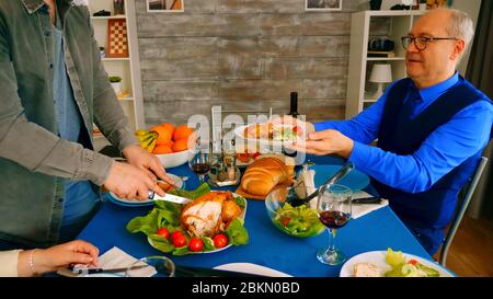 Zoom in shot di uomo che serve suo padre con pollo a cena di famiglia. Foto Stock