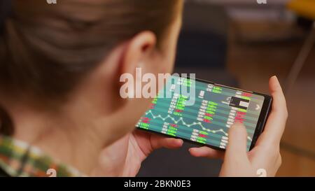 Vista posteriore della donna che controlla la negoziazione di azioni utilizzando lo smartphone. Foto Stock