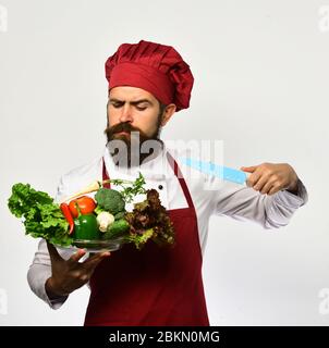 Uomo con barba isolata su sfondo bianco. Cuoco con faccia seria in uniforme di borgogna contiene gli ingredienti dell'insalata e coltello. Lo chef taglia verdure fresche. Cucina e dieta vegetariana Foto Stock