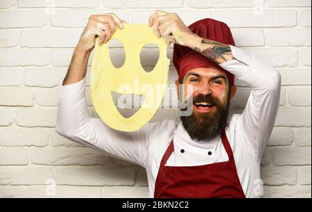 Uomo con barba tiene l'impasto laminato con smiley tagliato su sfondo bianco muro di mattoni. Lo chef prepara l'impasto. Cuoco con il viso eccitato in uniforme di borgogna gioca con l'impasto della pizza. Cucina italiana. Foto Stock