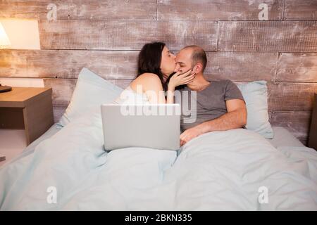 Bella moglie in pajama baciare il suo bel marito prima di andare a dormire. Laptop sul letto. Foto Stock