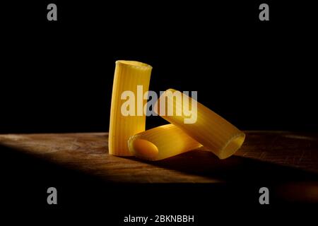Rigatoni, pasta tipica italiana disposta su un tavolo di legno attraversato da un fascio di luce su sfondo scuro Foto Stock