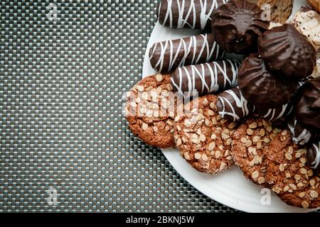 Un piatto bianco con biscotti d'avena, cosparsi di noci, marshmallows al cioccolato e dolci al cioccolato con strisce bianche su sfondo grigio-blu Foto Stock