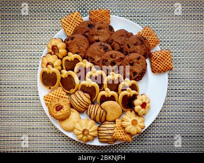 Biscotti alla farinata d'avena, biscotti al cioccolato in forma di mele e biscotti di forme diverse adagiati su un piatto bianco sullo sfondo di un wicke grigio Foto Stock