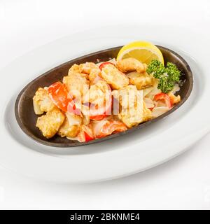 Fette di pesce fritto con pomodori, limone e prezzemolo adagiate in una padella su un piatto bianco Foto Stock