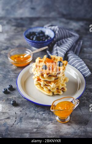 Tradizionali waffle belgi fatti in casa con mirtillo, miele e marmellata d'arancia su piastra su sfondo grigio. Foto Stock