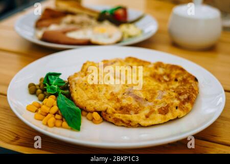 Frittata con prosciutto, piselli e mais in un bar. Deliziosa colazione con omelette, formaggio e piselli. Frittata con verdure su un piatto bianco in un accogliente caffè Foto Stock