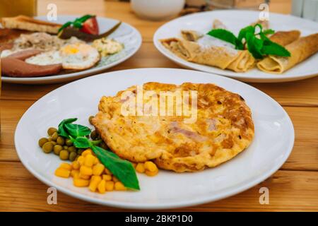 Frittata con prosciutto, piselli e mais in un bar. Deliziosa colazione con omelette, formaggio e piselli. Frittata con verdure su un piatto bianco in un accogliente caffè Foto Stock