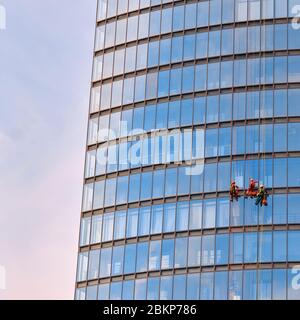 Tre uomini lavoratori in abiti da lavoro rosso e scuro pulizia delle finestre esterne di un grattacielo business, vista dal basso Foto Stock