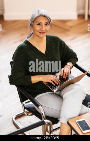 Foto di una donna felice dai capelli grigi che batte sul laptop e sorridente mentre si siede al tavolo in soggiorno Foto Stock