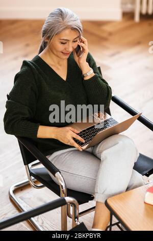 Foto di una donna soddisfatta dai capelli grigi che usa il laptop e parla al cellulare mentre si siede al tavolo in soggiorno Foto Stock