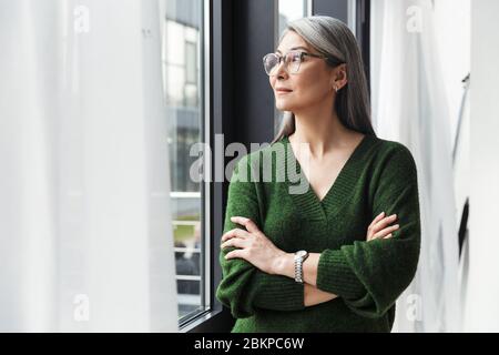 Donna matura sorridente attraente con lunghi capelli grigi in piedi alla finestra al chiuso Foto Stock
