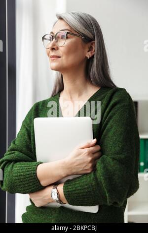 Foto di una donna d'affari seria dai capelli grigi in occhiali che tiene il laptop mentre lavora in ufficio Foto Stock