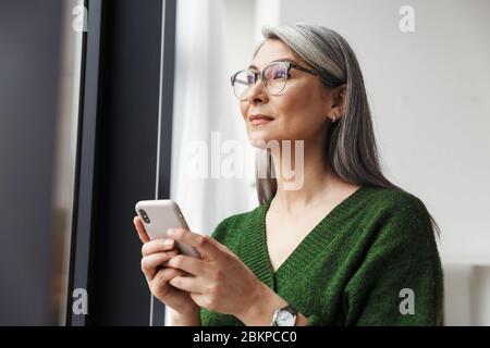Foto di grigio-capelli donna d'affari seria in occhiali utilizzando cellulare mentre lavorano in ufficio Foto Stock
