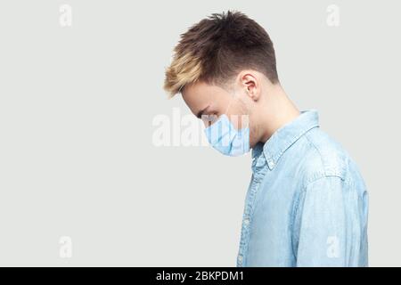 Profilo vista laterale ritratto di triste rovesciamento solo giovane uomo con maschera chirurgica medica in camicia blu in piedi, tenendo testa giù e depresso. Stu indoor Foto Stock